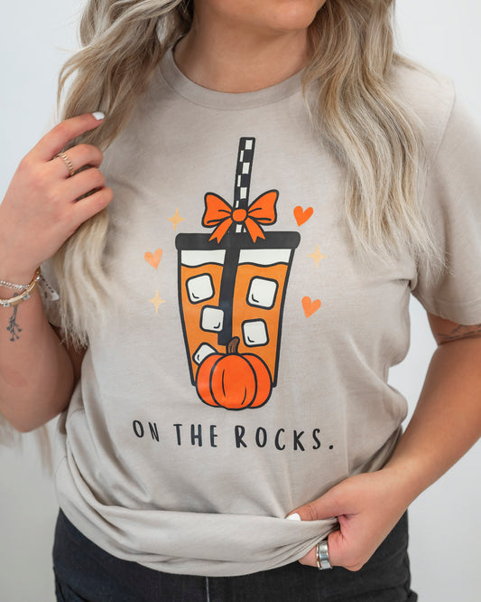Person wearing a beige t-shirt with a graphic design of a pumpkin drink and text 'On the Rocks.'