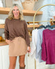 Woman wearing a brown sweater and beige skirt standing in front of a clothing rack with various garments.