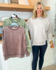 Woman standing next to two sweaters, one green and one brown, in a home setting.