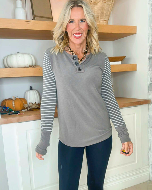 Woman wearing a gray long-sleeve top with striped sleeves and blue leggings in a room with wooden shelves and pumpkins.