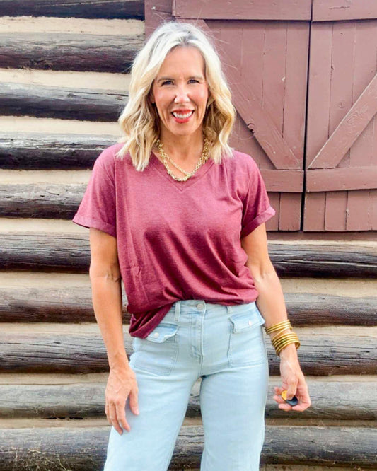 Woman wearing a maroon t-shirt and light blue jeans standing in front of a wooden wall.