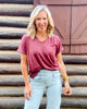 Woman wearing a maroon t-shirt and light blue jeans standing in front of a wooden wall.