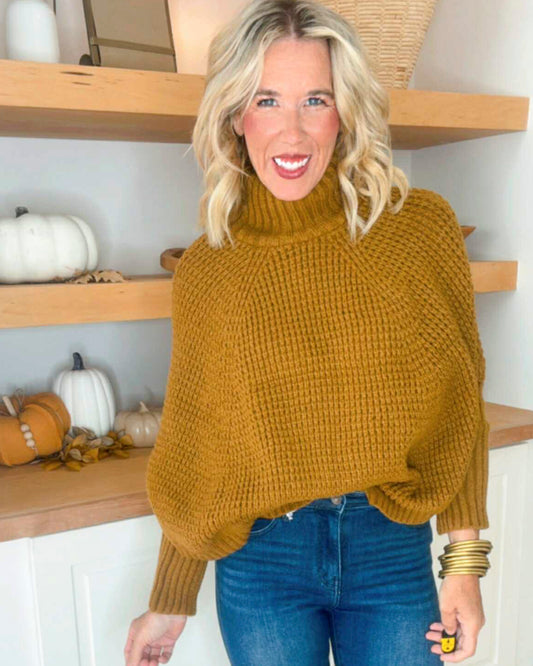 Woman wearing a mustard yellow sweater in a home setting with wooden shelves and pumpkins.