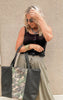 Woman wearing olive wide-leg pants and black tank holding a two-toned camo and black weekender tote from the Makeup Junkie x Bad Habit collection.