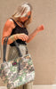 Woman wearing a black tank and olive pants holding the camo Makeup Junkie Daykeeper Tote with tassel detail.
